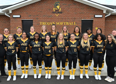 Team photo of the softball team in black and gold uniforms standing in front of the brick fieldhouse.