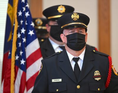 Firefighters in dress uniform walk in a line carrying the USA flag.