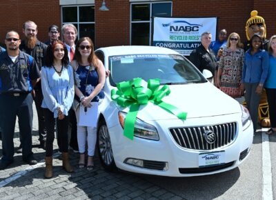 A group of people stand around a white car with a large green ribbon on the hood.