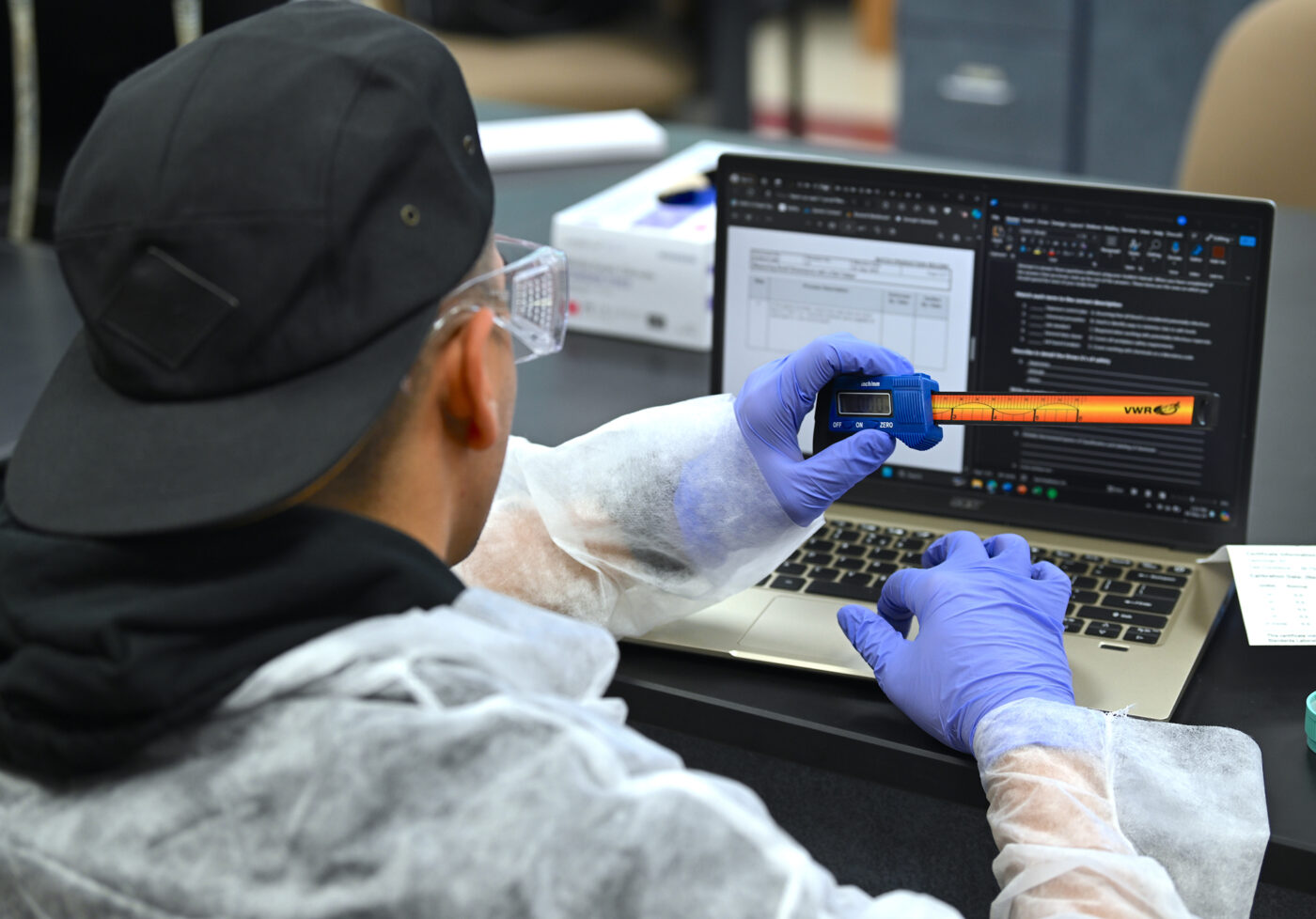 A student sits a computer and holds a caliper for measuring.