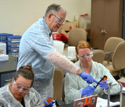 A man in a lab coat and safety glasses points to a measuring instrument held by a woman seated at a lab table.