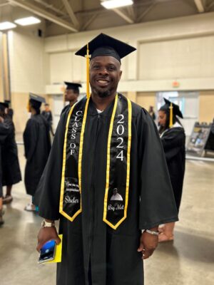 Emond Virgil stands wearing a graduation cap and gown, as well as a black stole with gold trim.