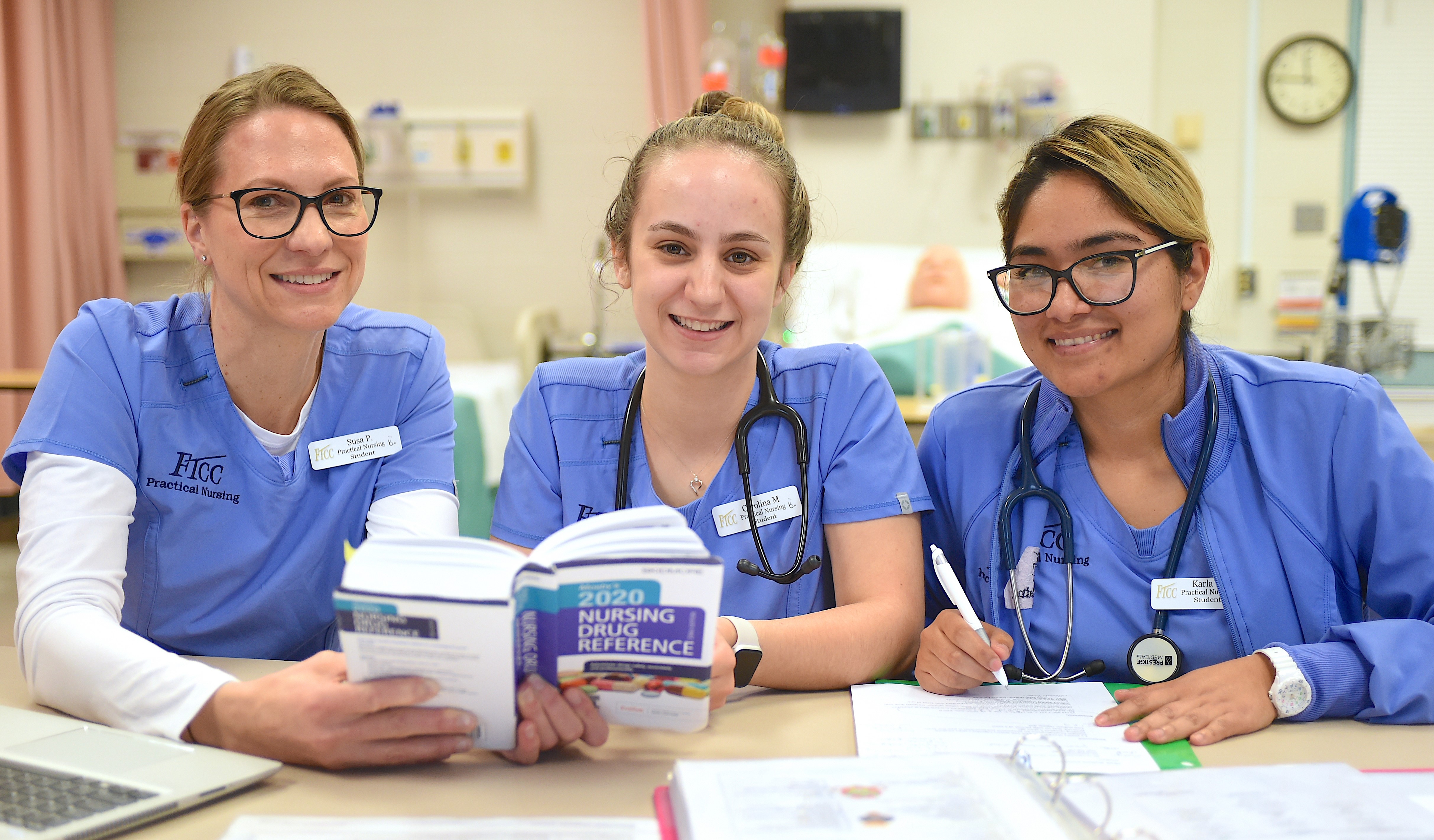Three nursing students in blue scrubs studying together at a table with books and notes in a classroom or lab setting.
