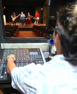 A student adjusts settings on a sound board while musicians perform on stage.