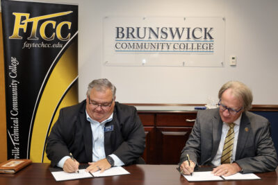 Two men in suits sign paperwork while sitting at a table.