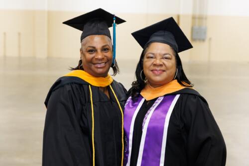 Two instructors, dressed in graduation regalia, pose for a photo.