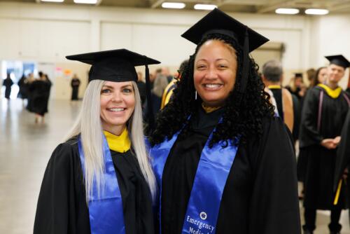 Two graduates dressed in caps and gowns with blue stoles pose for a photo.