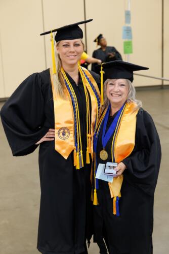 Two graduates with gold stoles pose for a photo. Both graduates are wearing several cords, and the graduate on the right has a medallion on a blue cord.