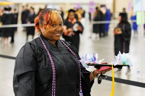 A graduate in a gown with a red, white and blue cord smiles at the camera while holding her cap.