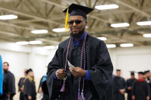 A graduate dressed in cap and gown and wearing sunglasses walks through the staging area.