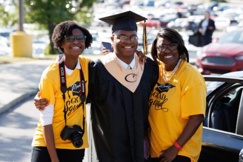 A graduate wearing his cap and gown and a gold stole with a hawk logo on it stands with his family.