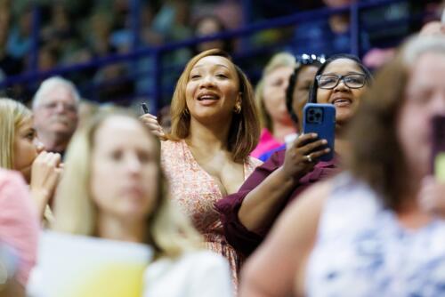 Two women standing in the audience smile as they look at the graduates.