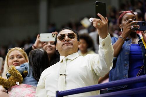 A man wearing sunglasses and a necktie and dress shirt holds his phone up.