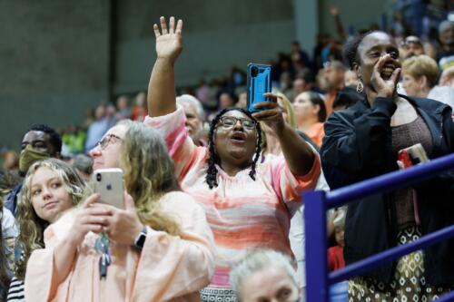 A woman raises her hand and cheers while holding her phone up in the stands.