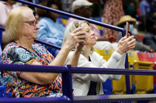 Two women, shown in profile, hold up their phones while seated in the stands.