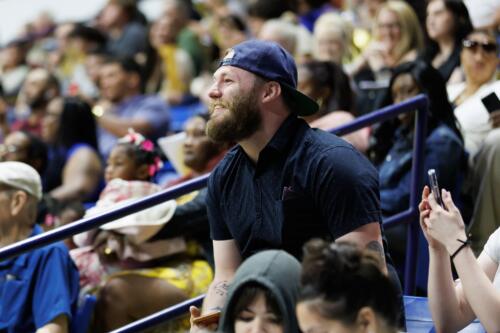 A bearded man wearing a backwards ball cap smiles as he leans forward in his seat.