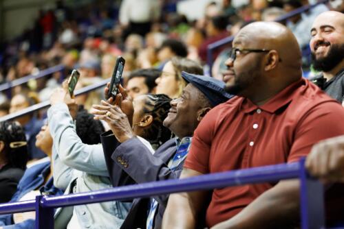 A man seated in a group smiles as he holds up his phone to take a photo from his seat.