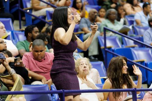 A woman, shown in profile, raises her hands as she cheers in the stands.