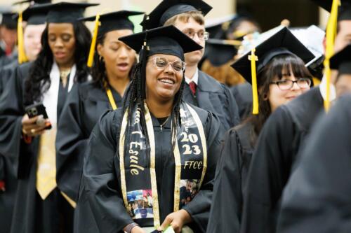 A graduate wearing a black and gold personalized stole waits in line in the staging area.