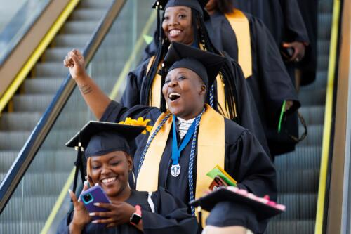 Graduates descend the escalator. One of them, wearing a gold stole and a medallion on a blue ribbon, raises a hand and cheers.