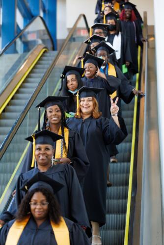 Graduates descend the escalator. One of them holds up two fingers in a "peace sign."