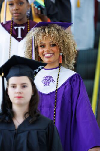 A graduate in a purple gown with a white stole with a knight's helmet on it smiles at the camera.