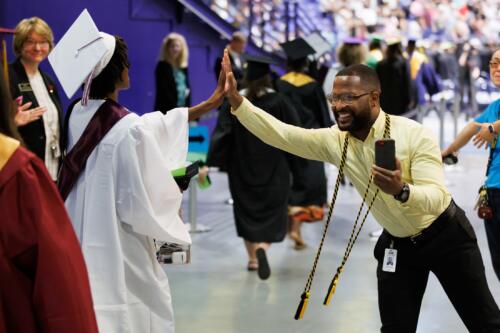 A graduate in a white cap and gown high fives Gary Womble, who is dressed in a yellow shirt and black pants.