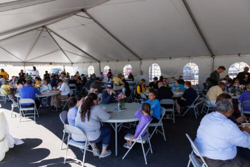 A wide photo of people sitting down to eat lunch at several tables inside a tent.