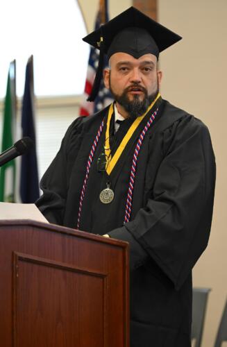 A male graduate wearing a cap, gown medal and cord speaks at the podium. 