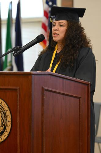 A female graduate wearing a cap, gown and medal around her neck speaks at the podium.