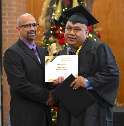 A graduate poses with his certificate while standing next to Miguel Figueroa.