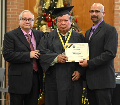 A graduate poses with his certificate while standing next to Miguel Figueroa and another man in a suit.