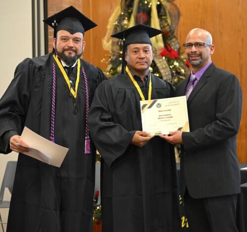 A graduate poses with his certificate while standing next to Miguel Figueroa and his fellow graduate.