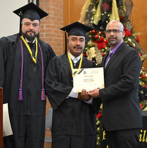 A graduate poses with his certificate while standing next to Miguel Figueroa and his fellow graduate.