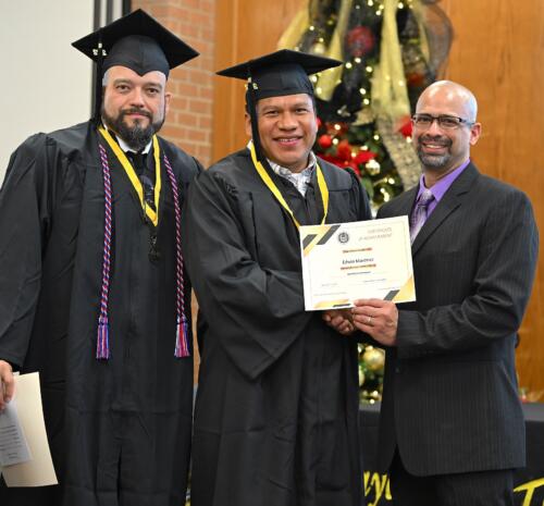 A graduate poses with his certificate while standing next to Miguel Figueroa and his fellow graduate.