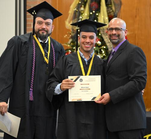 A graduate poses with his certificate while standing next to Miguel Figueroa and his fellow graduate.