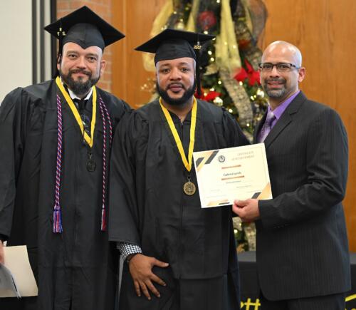 A graduate poses with his certificate while standing next to Miguel Figueroa and his fellow graduate.