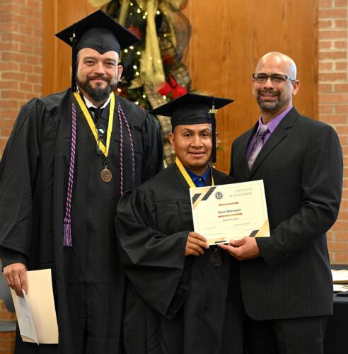 A graduate poses with his certificate while standing next to Miguel Figueroa and his fellow graduate.