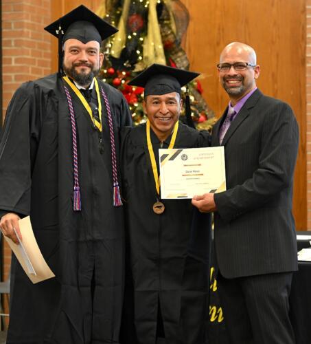 A graduate poses with his certificate while standing next to Miguel Figueroa and his fellow graduate.