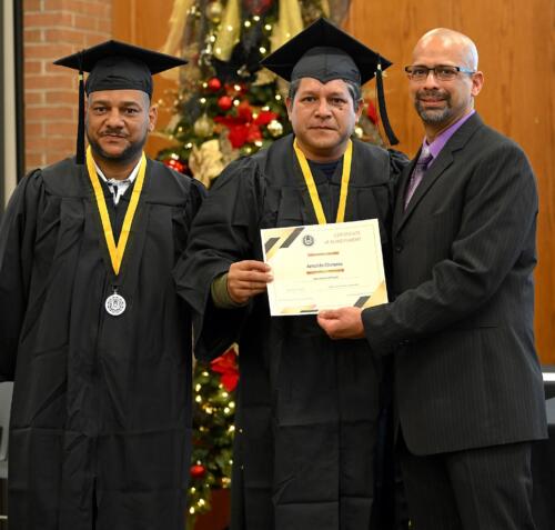 A graduate poses with his certificate while standing next to Miguel Figueroa and his fellow graduate.