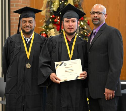 A graduate poses with his certificate while standing next to Miguel Figueroa and his fellow graduate.