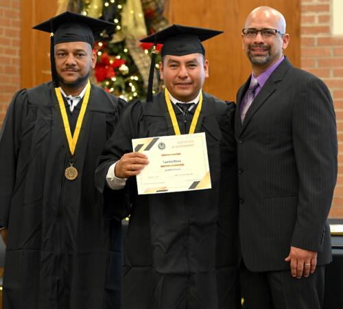 A graduate poses with his certificate while standing next to Miguel Figueroa and his fellow graduate.